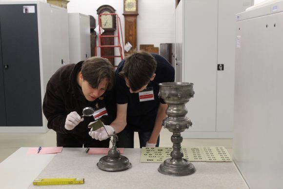 Zwei junge M&auml;nner beugen sich &uuml;ber eine Pokalfigur im Depot des Museums. Sie tragen wei&szlig;e Handschuhe, ein Junge h&auml;lt eine Lupe in der Hand und schaut durch