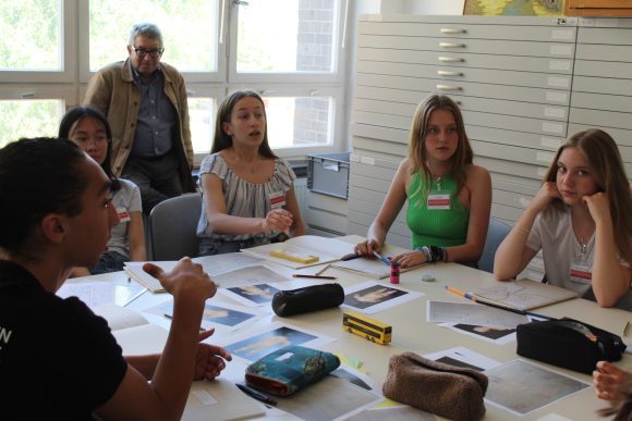 Eine Gruppe von Jugendlichen sitzt um einen Tisch, darauf sind Bl&ouml;cke, Stifte und Papier zu sehen.